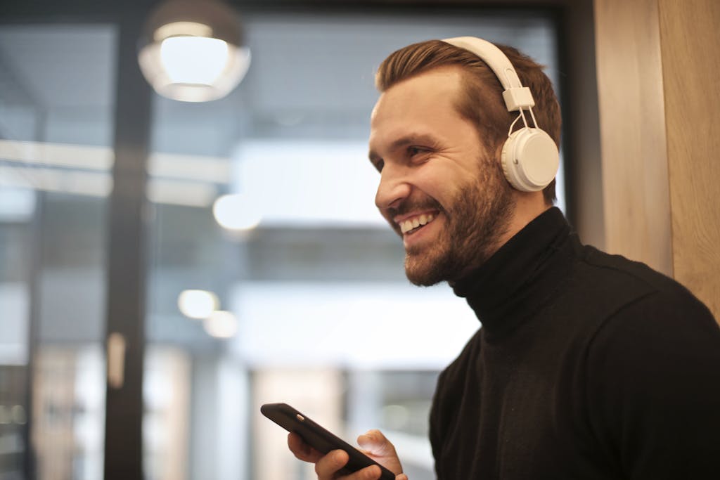 Bearded man smiling with headphones and smartphone indoors, enjoying music.